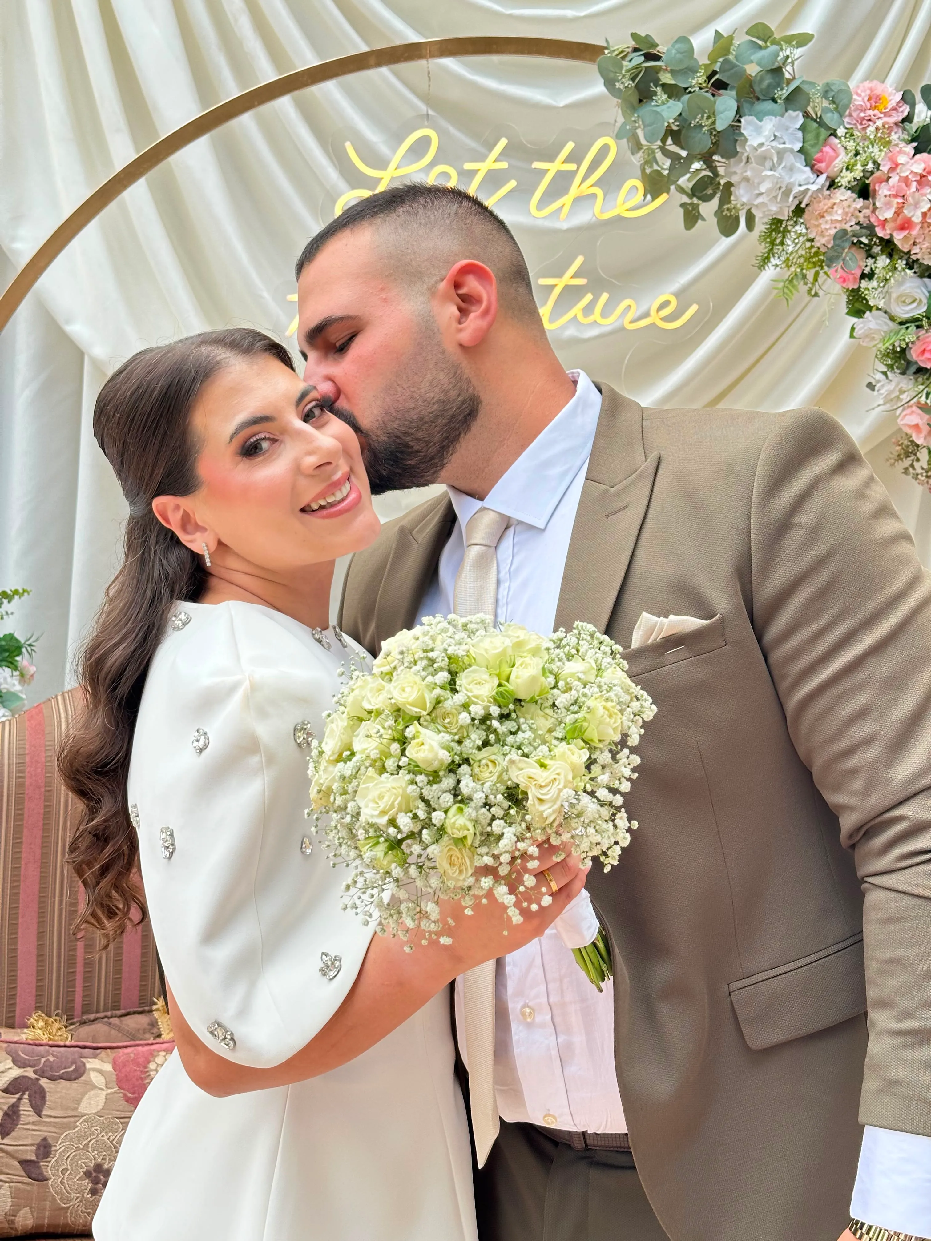 The groom kissing the bride's cheek under a flower arch, the bride smiling with a bouquet of white roses